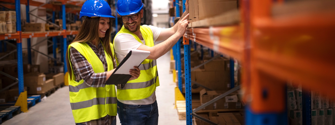 A female and male employee wearing yellow construction vests and blue hard hats landing in a warehouse and looking at a notepad.