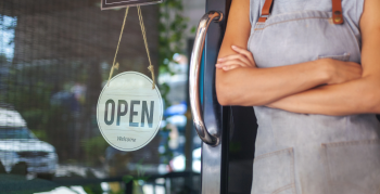 Shop worker next to a storefront&#039;s &quot;Open&quot; sign.