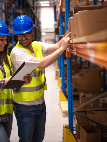 A female and male employee wearing yellow construction vests and blue hard hats landing in a warehouse and looking at a notepad.